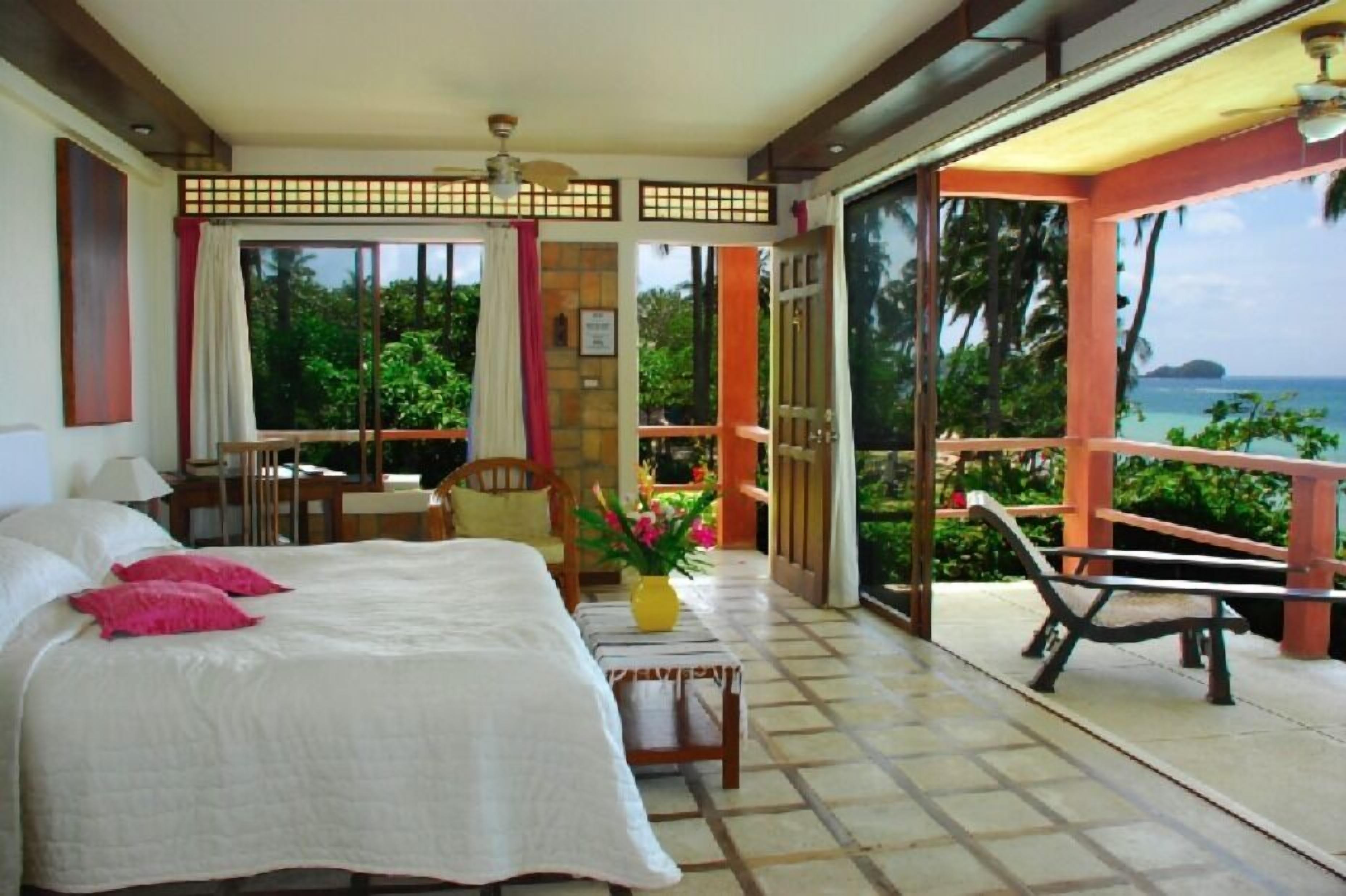 Quarto de hotel com cama, uma mesinha com um vaso, uma cadeira e varanda com vista para o mar.