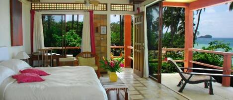 Quarto de hotel com cama, uma mesinha com um vaso, uma cadeira e varanda com vista para o mar.
