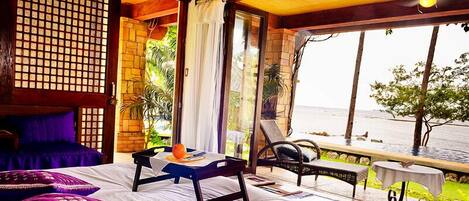 A room with a bed, a table with a fruit and a book, a chair, and a view of the sea.
