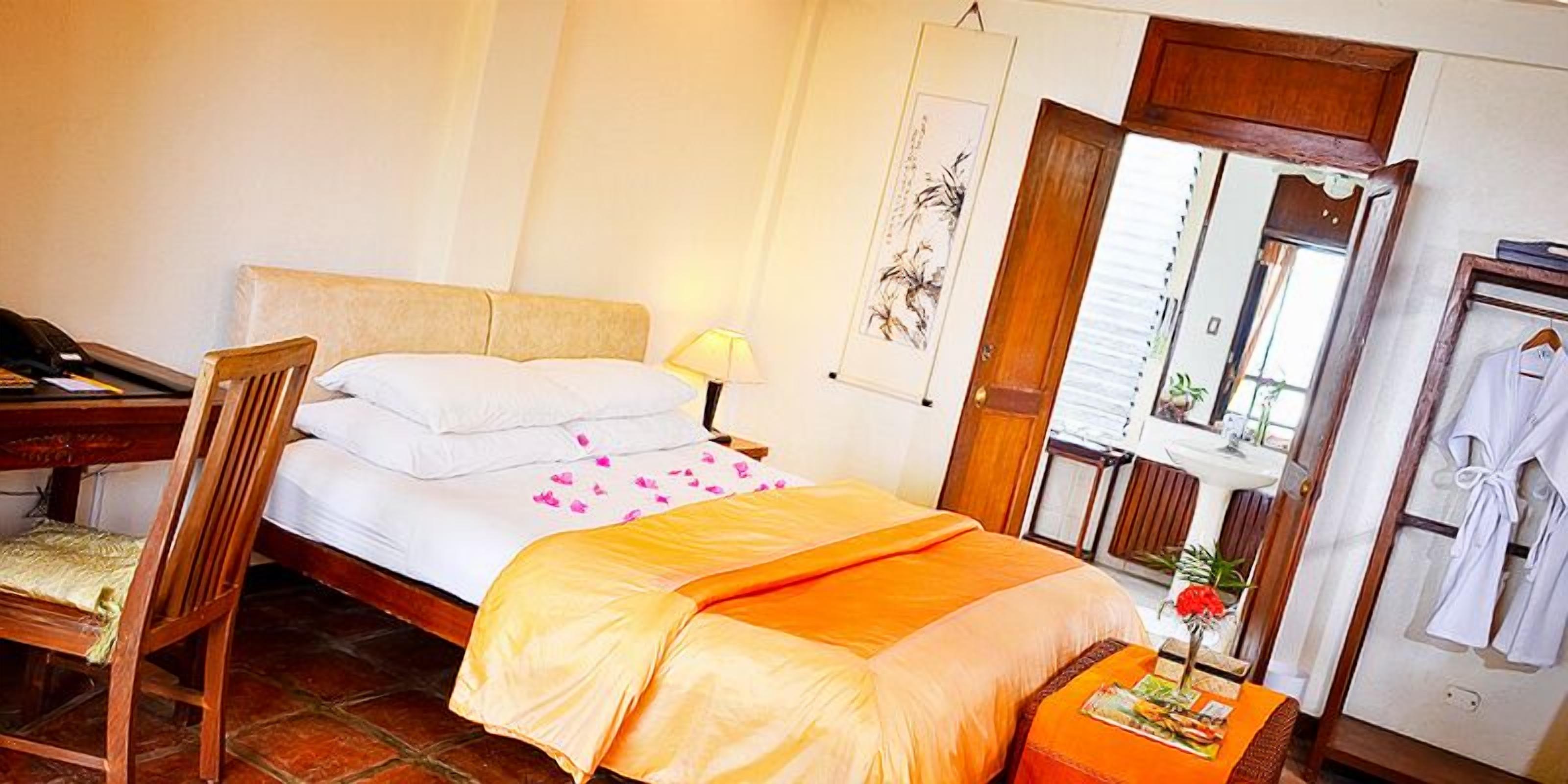 Habitación de hotel con cama, escritorio, silla, mesita de noche con lámpara, espejo y bata colgada en la pared.