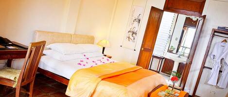 Habitación de hotel con cama, escritorio, silla, mesita de noche con lámpara, espejo y bata colgada en la pared.