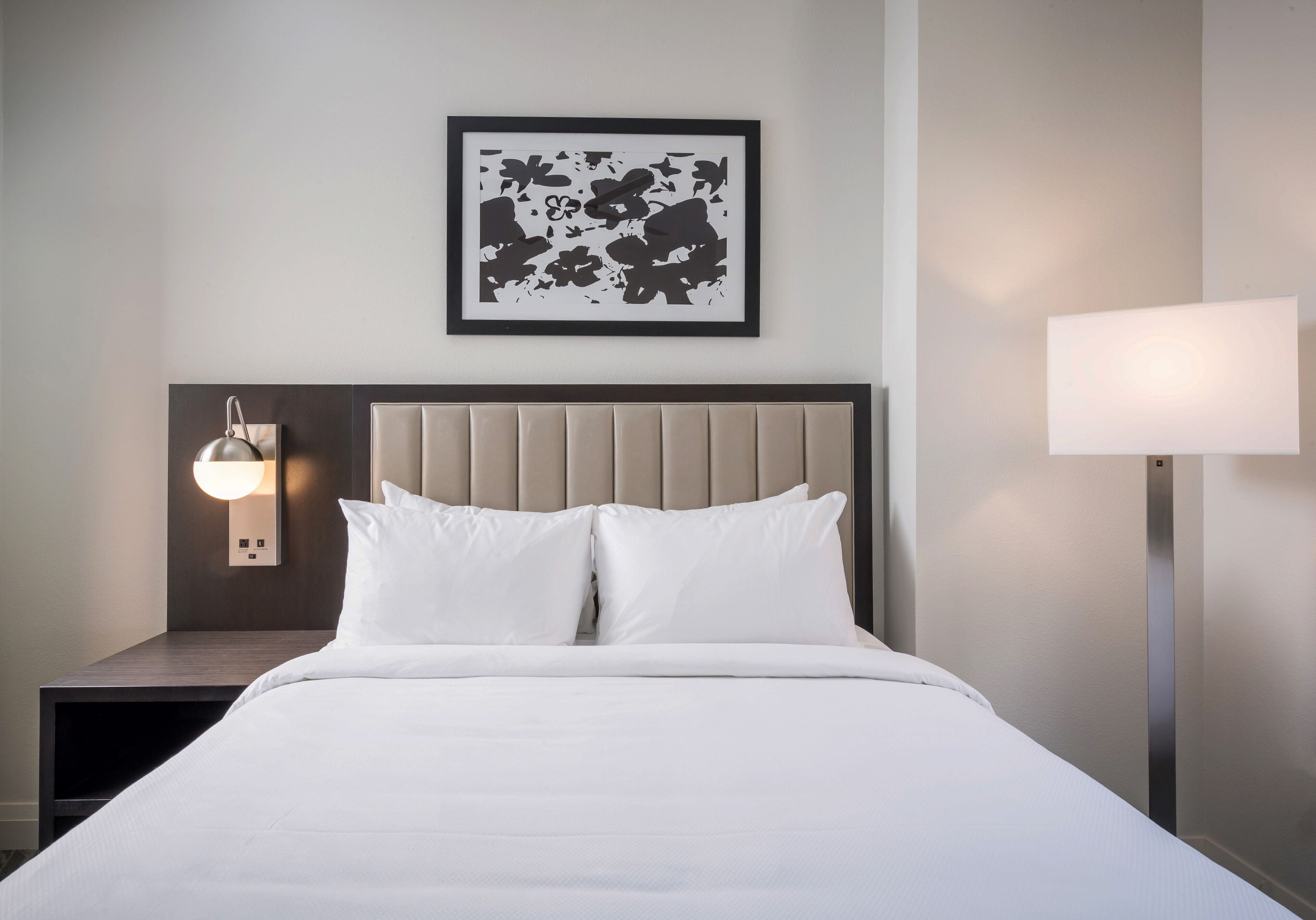 A hotel room with a bed, a nightstand, a wall-mounted lamp, and a framed artwork above the bed.