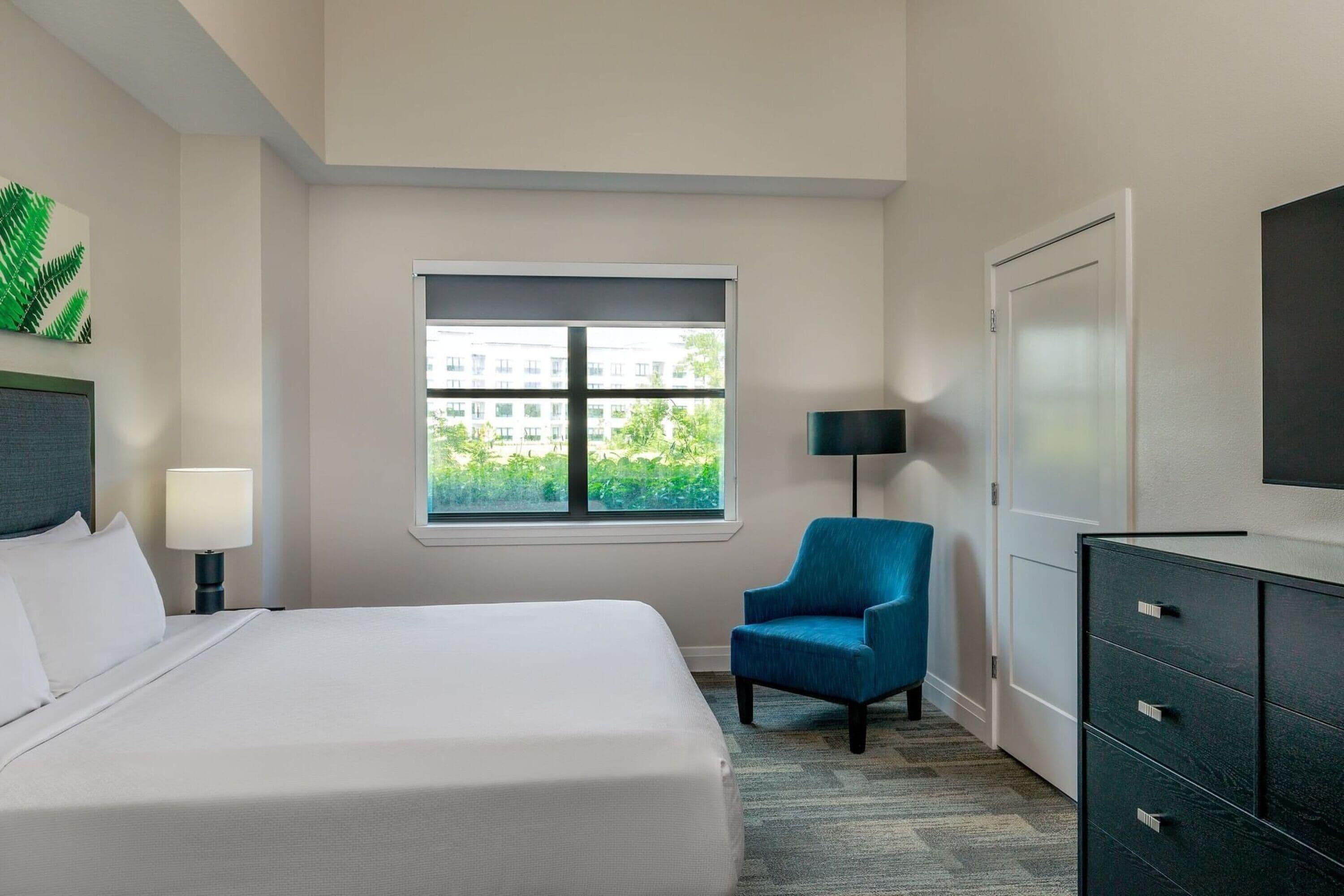 A hotel room with a bed, a chair, a desk, a lamp, a TV, and a window with a view of buildings and greenery.