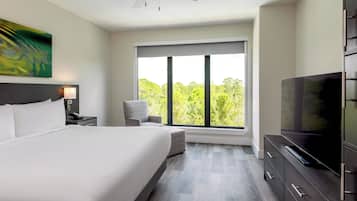 A modern hotel room with a large bed, a TV, a desk with a lamp, and a window with a view of trees.