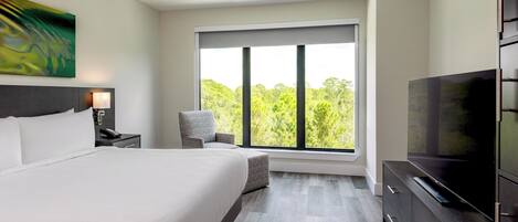 A modern hotel room with a large bed, a TV, a desk with a lamp, and a window with a view of trees.