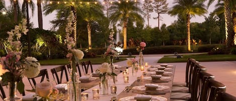 Un espacio de comedor al aire libre con luces colgantes, palmeras y centros de mesa florales.
