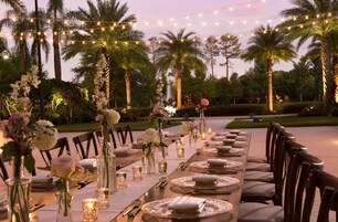 Un espacio de comedor al aire libre con luces colgantes, palmeras y centros de mesa florales.