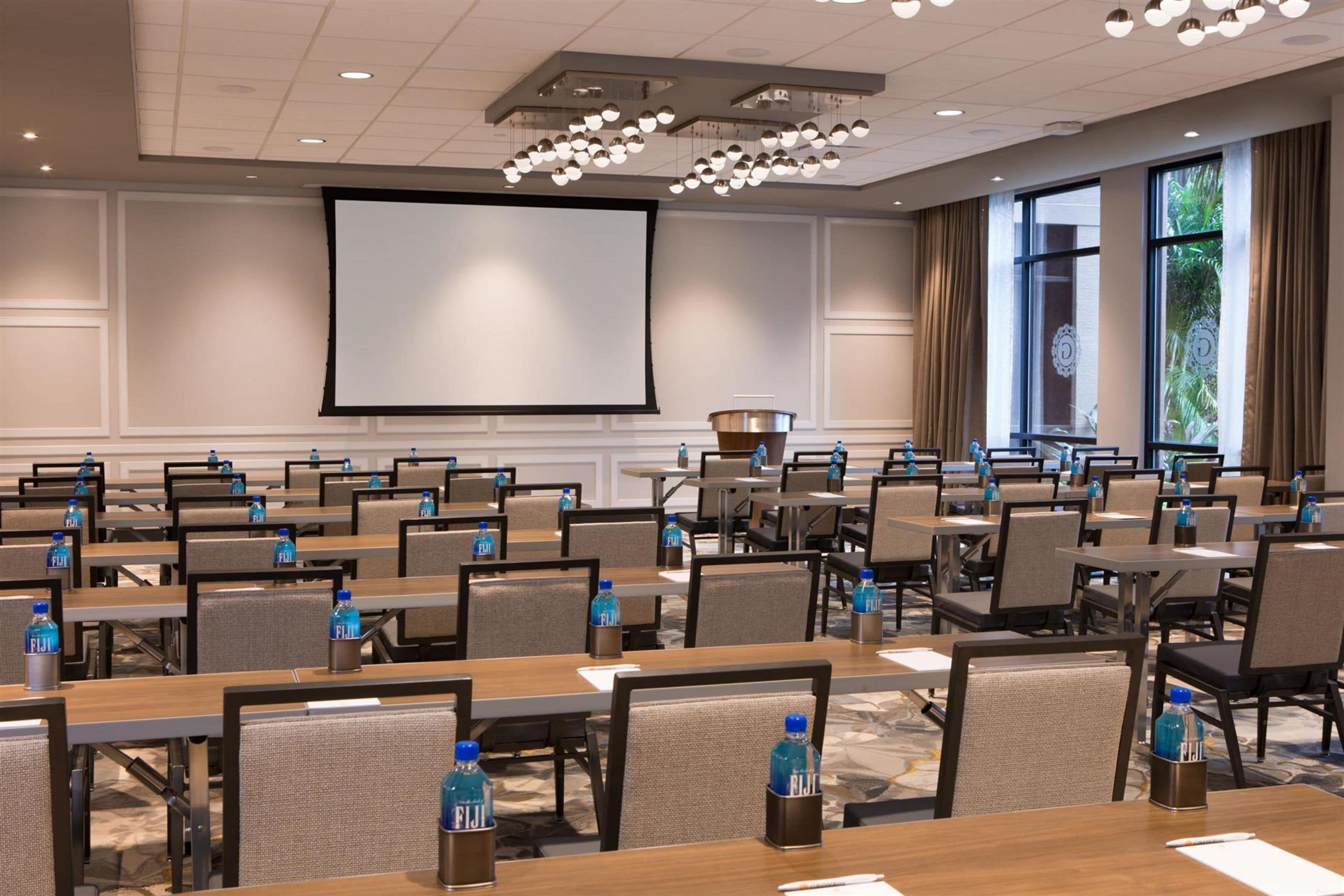 A conference room with rows of tables, chairs, and a large screen.