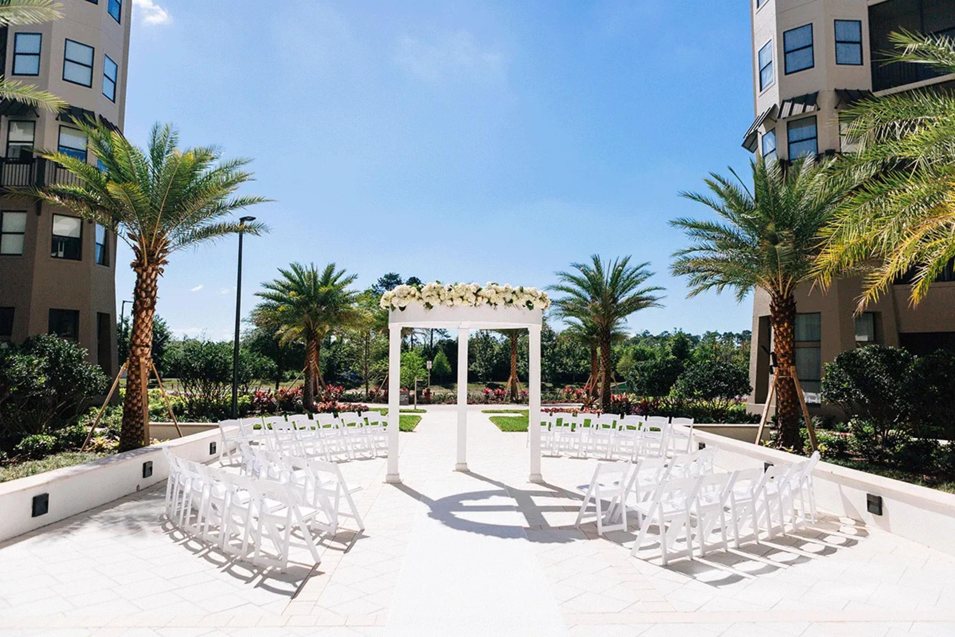 Un lieu de réception en plein air pour les mariages, avec des chaises blanches disposées en rangées, un gazebo central également blanc, agrémenté de décorations florales, et des palmiers.