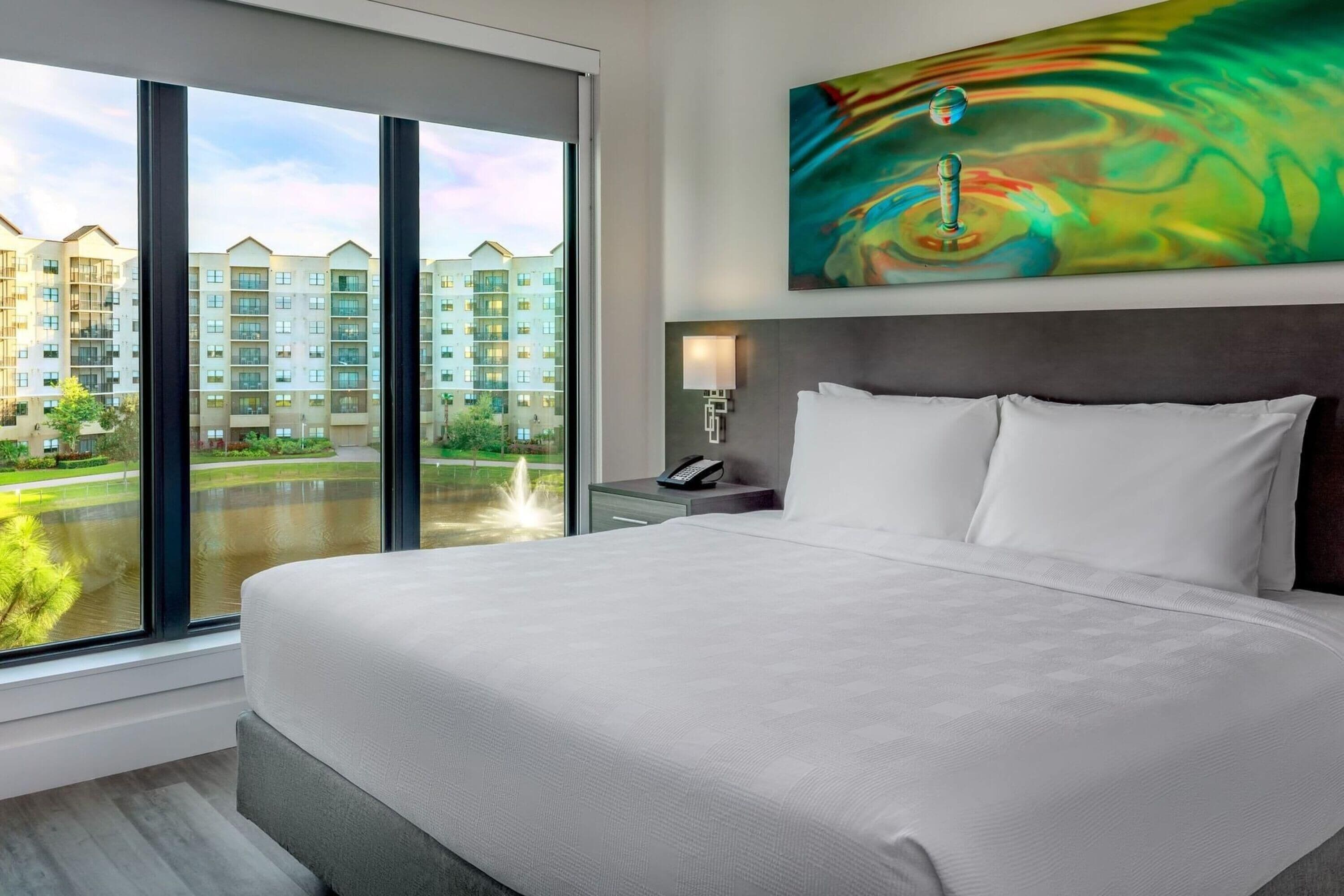 A hotel room with a large bed, a view of a building complex, and a colorful abstract painting on the wall.