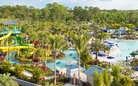 A resort with a large pool, water slides, palm trees, and beach umbrellas.