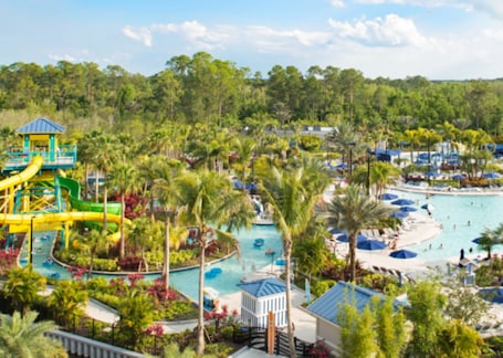 A resort with a large pool, water slides, palm trees, and beach umbrellas.
