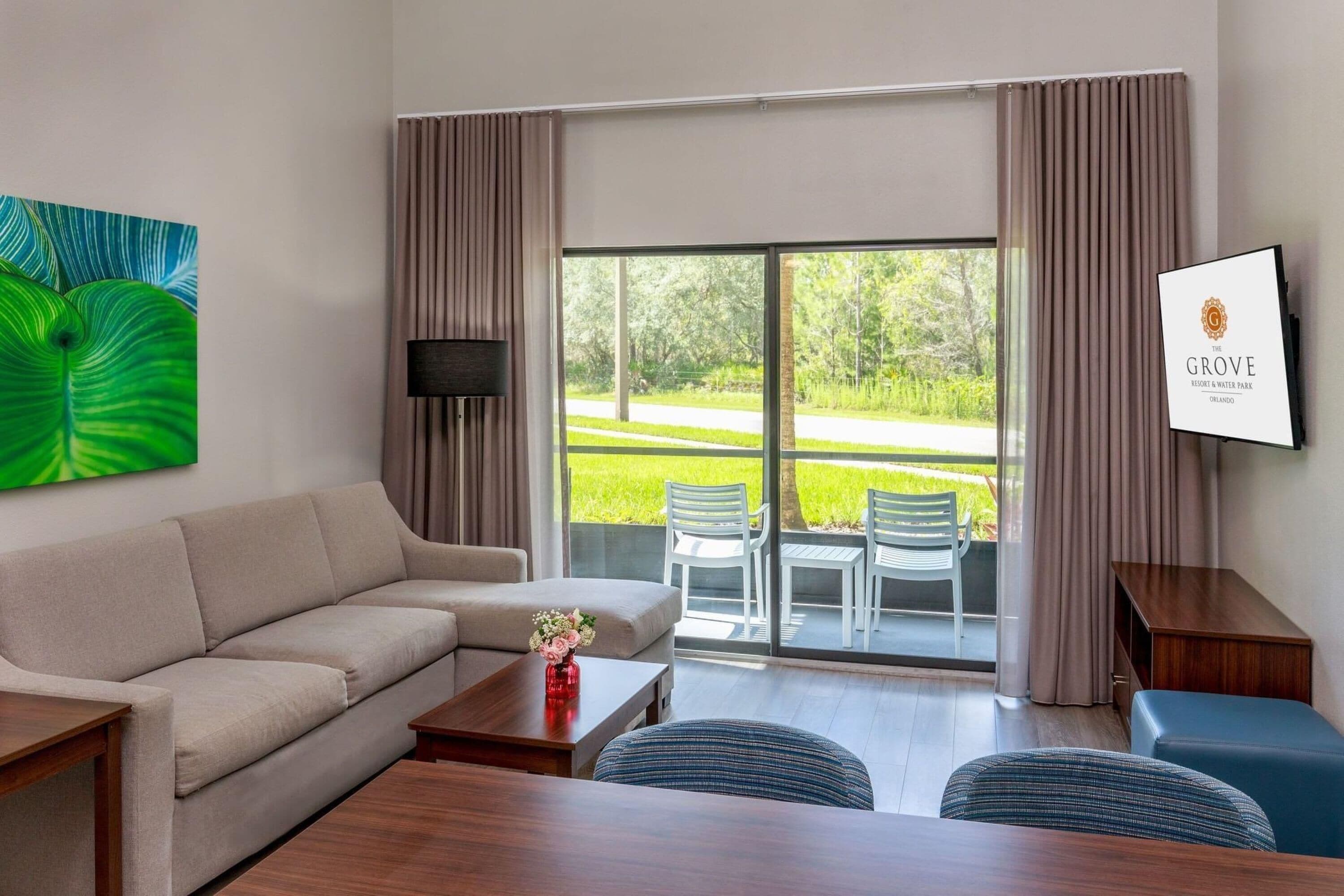 A hotel room with a sofa, a dining table, a TV, a large window with curtains, and a sign indicating the hotel name.