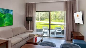Habitación de hotel con sofá, mesa de comedor, televisor, ventana grande con cortinas y un cartel con el nombre del hotel.