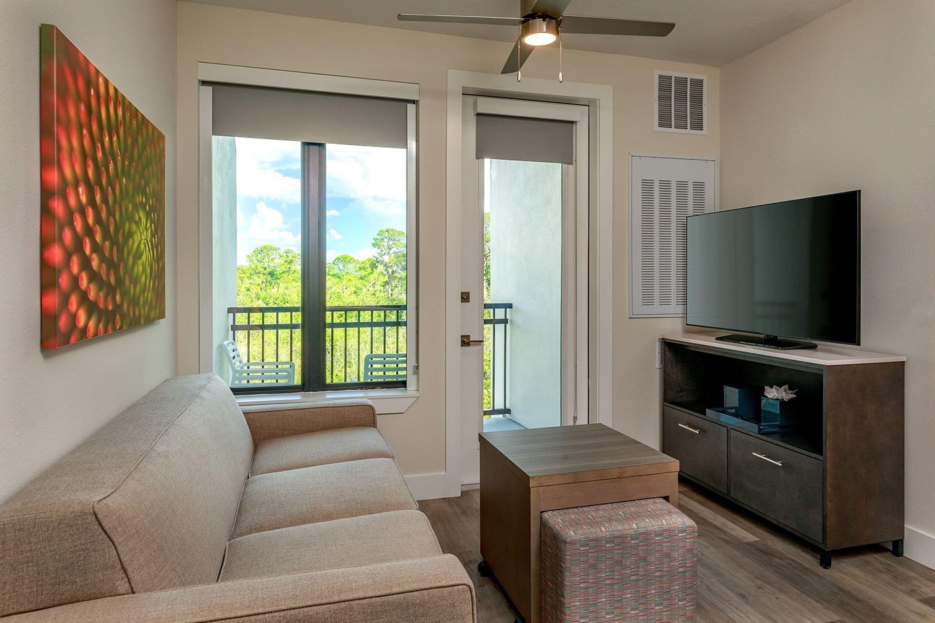 A living room with a sofa, a television on a cabinet, a ceiling fan, and a sliding glass door leading to a balcony.