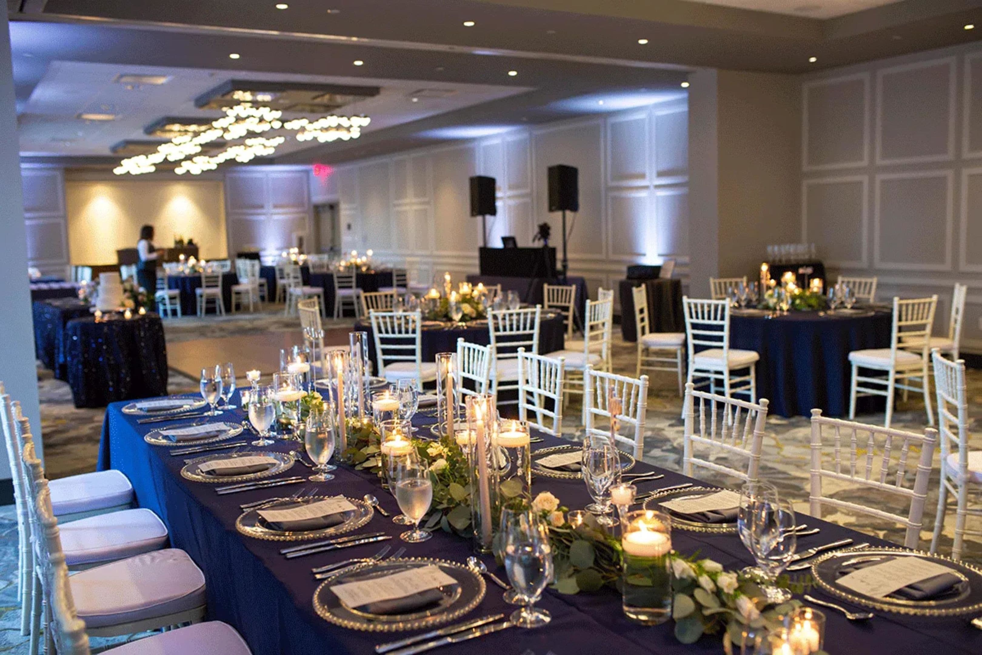 A banquet hall with round tables set for a formal event, featuring white chairs, dark tablecloths, and floral centerpieces.