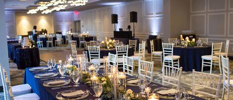 Une salle de banquet avec des tables rondes dressées pour un événement formel, agrémentée de chaises blanches, de nappes sombres et de centres de table floraux.