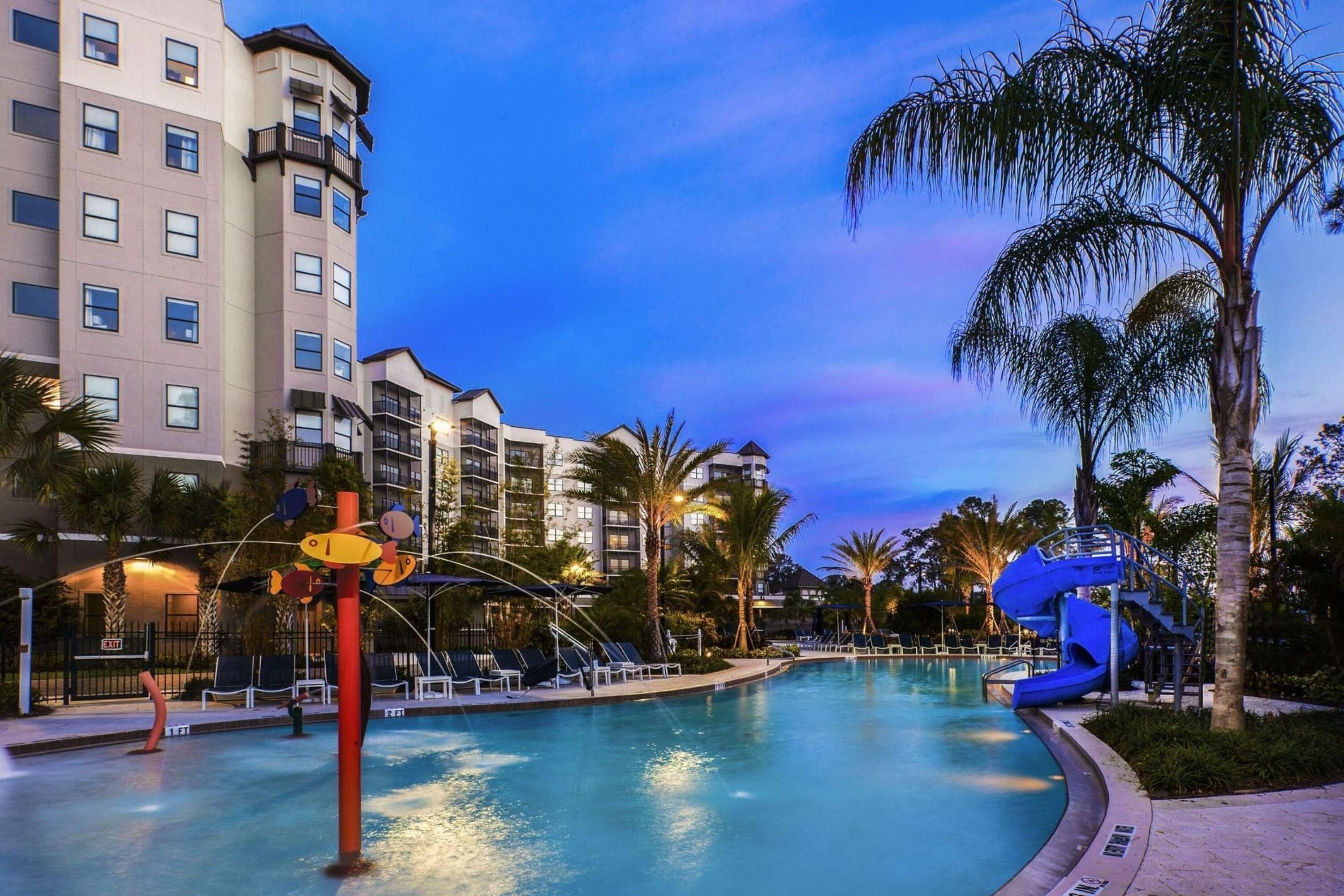A swimming pool with a slide, palm trees, and a building with balconies.