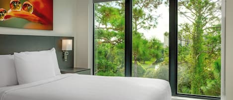 A hotel room with a large bed, a nightstand, a wall-mounted lamp, and a large window offering a view of trees and greenery.