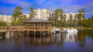 A wooden gazebo by a calm lake with a boat dock, surrounded by multi-story buildings.