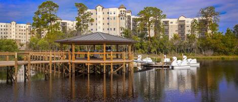 A wooden gazebo by a calm lake with a boat dock, surrounded by multi-story buildings.