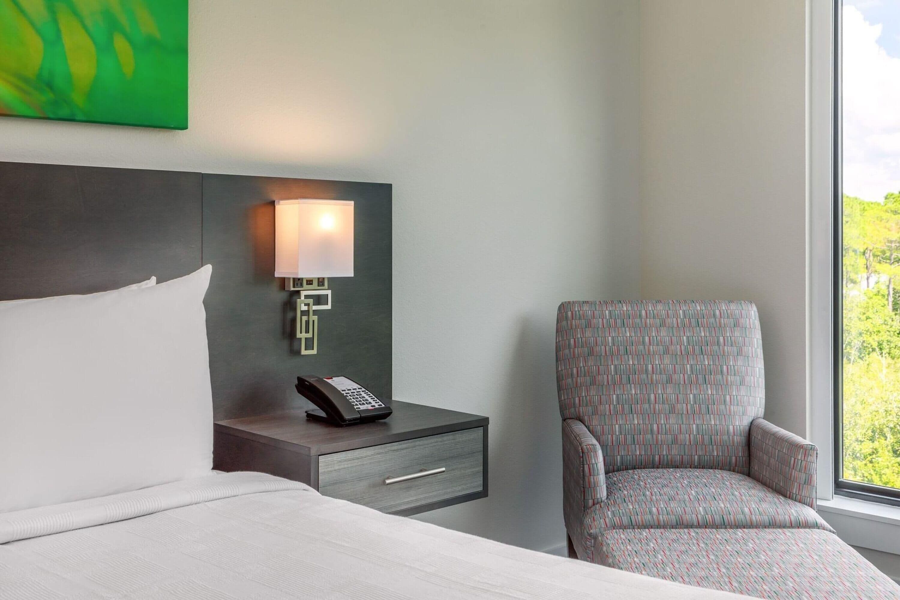 Une chambre d’hôtel avec un lit, une table de chevet avec une lampe, un téléphone et un fauteuil à motifs.