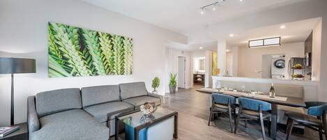 A modern living room with a grey sofa, a glass coffee table, and a dining area with blue chairs.