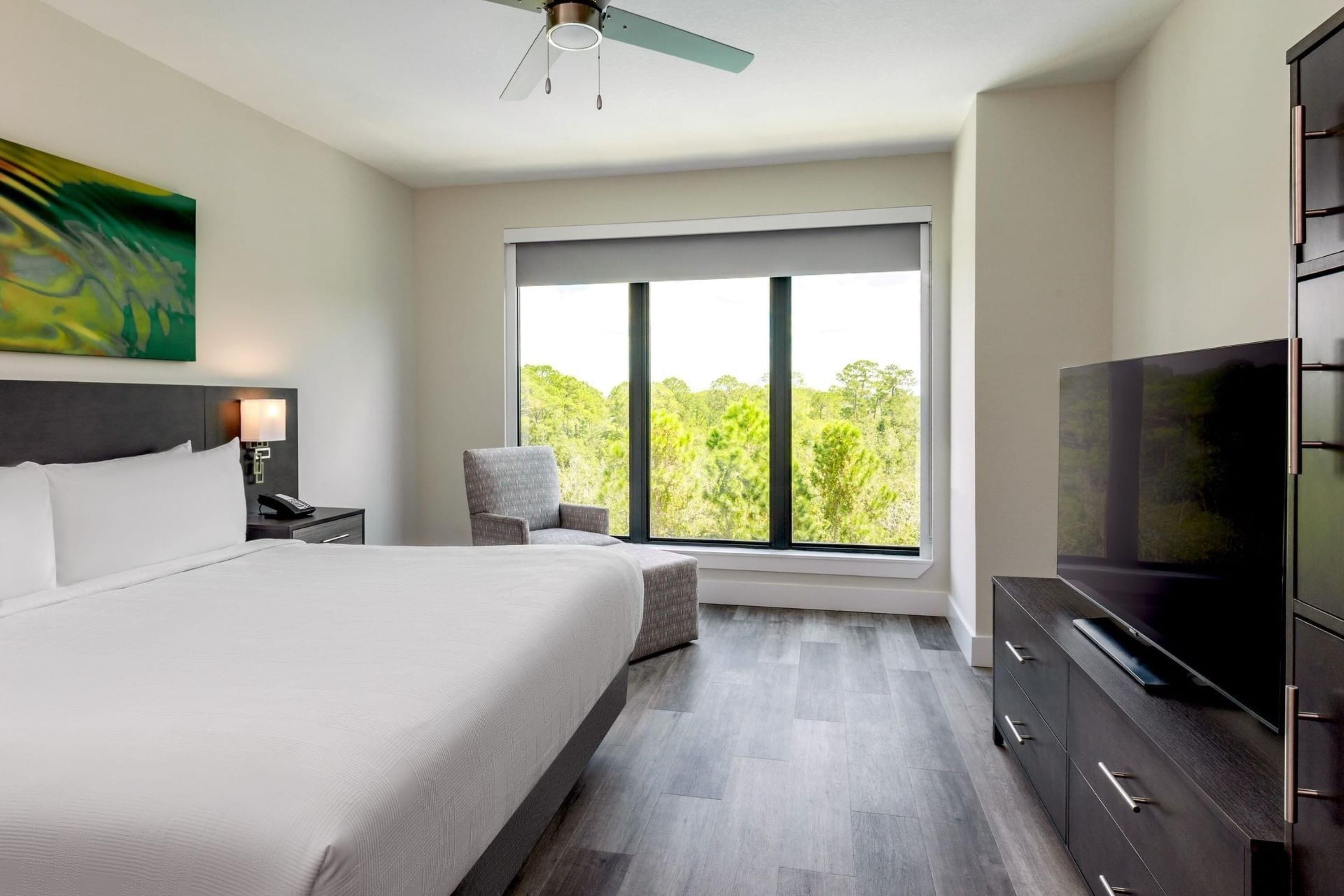 A modern hotel room with a large bed, a TV, a desk with a lamp, and a window with a view of trees.
