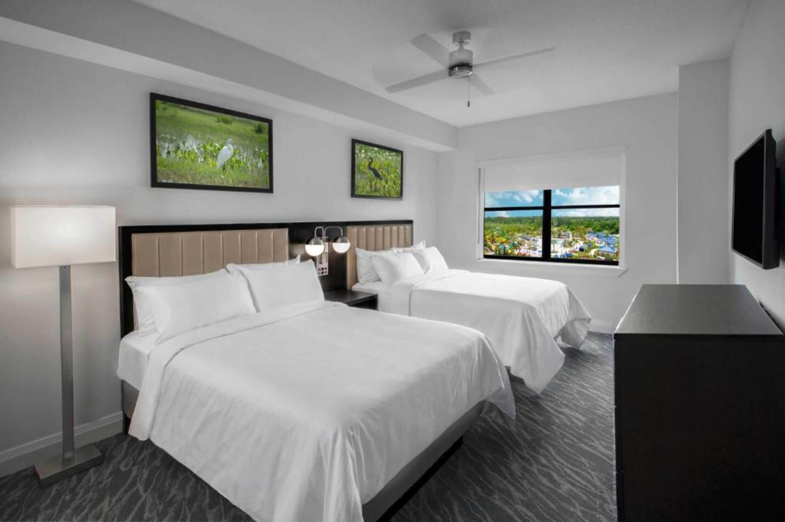 A hotel room with two beds, a flat-screen TV, a ceiling fan, and a window with a view of a landscape.