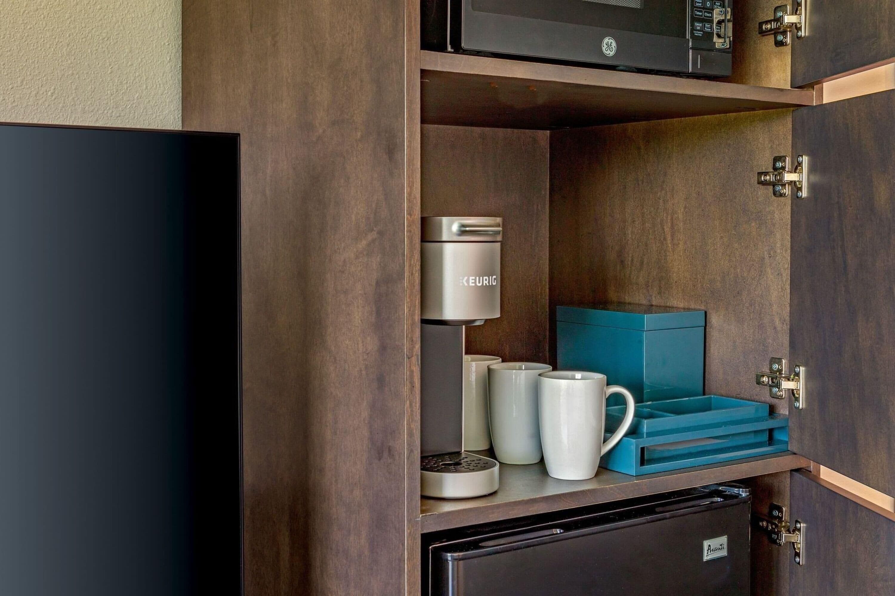 A kitchenette with a GE coffee maker, white mugs, a blue storage box, and a refrigerator.
