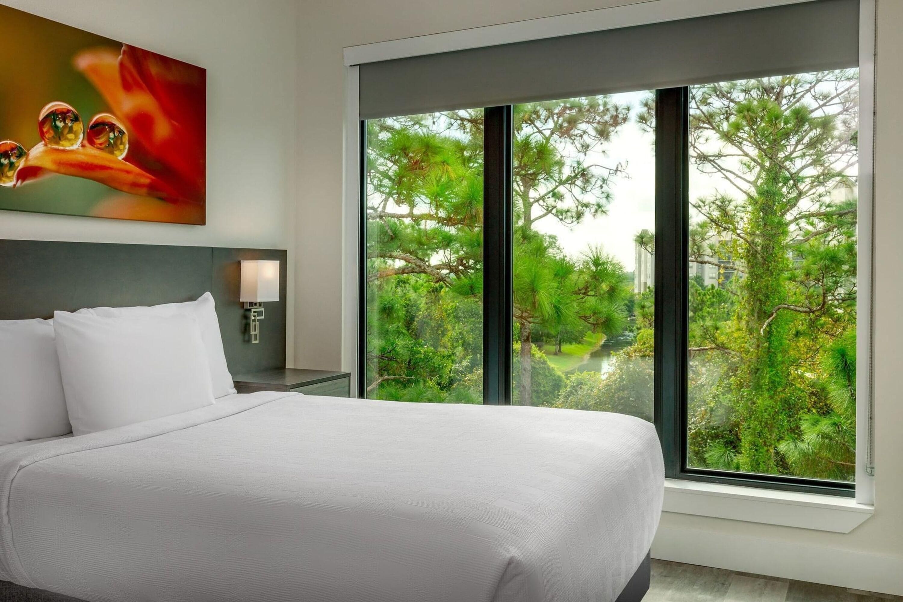 A modern hotel room with a large bed, a nightstand, a wall-mounted lamp, and a large window offering a view of trees and greenery.