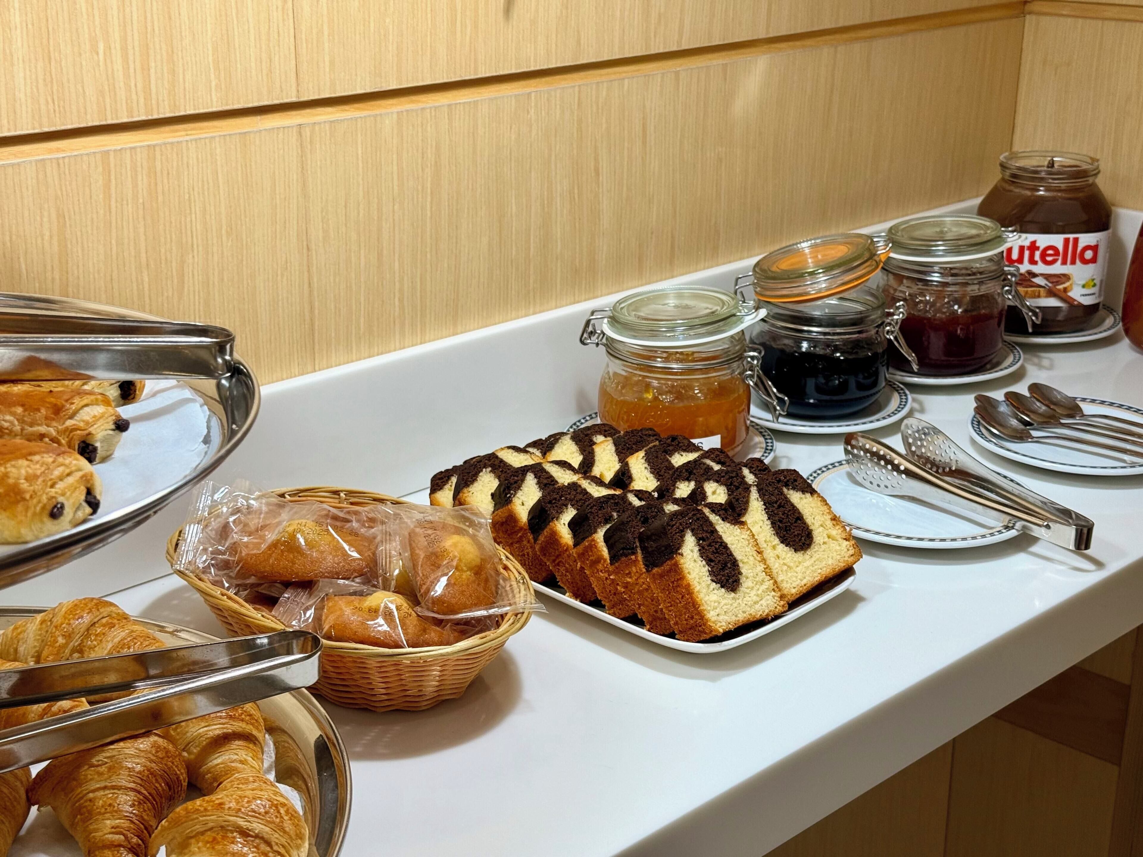A buffet table with various baked goods, including pastries and a cake, along with condiments like Nutella and jams.