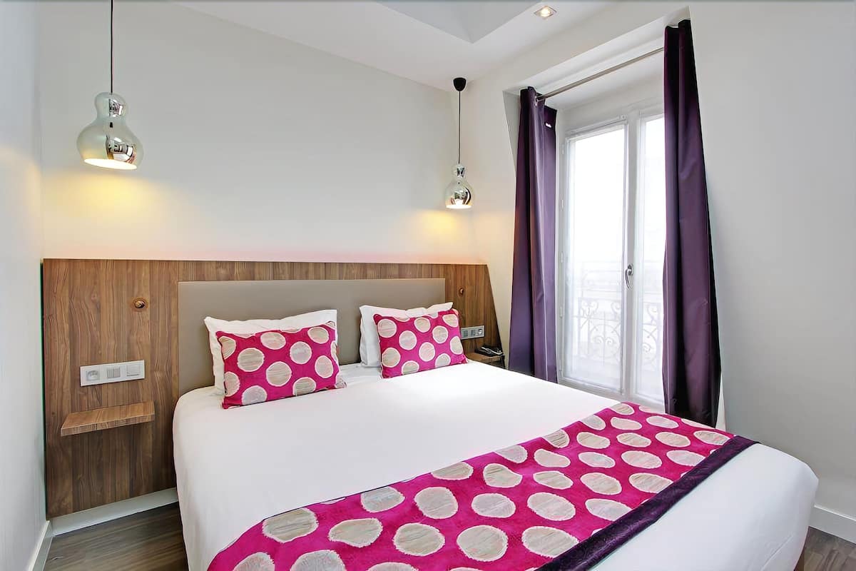 Quarto com cabeceira de madeira, cama branca com almofadas estampadas em rosa e branco, e cortina roxa.