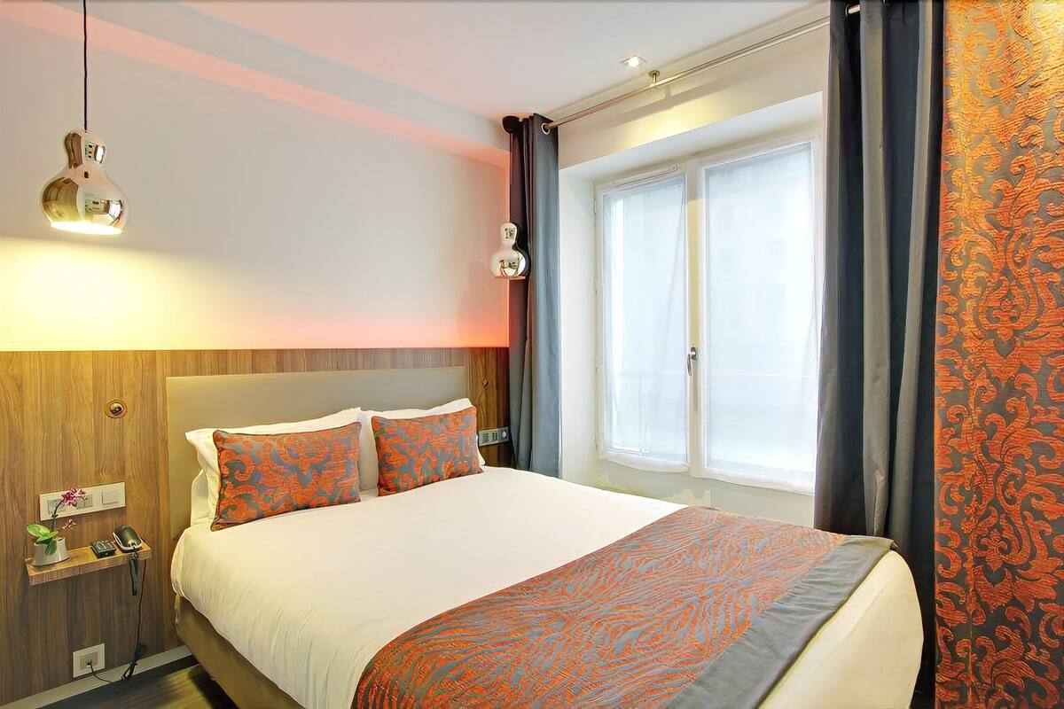 Quarto de hotel com uma cama grande, uma mesa de cabeceira com telefone e planta, uma luminária na parede e uma janela com cortinas.
