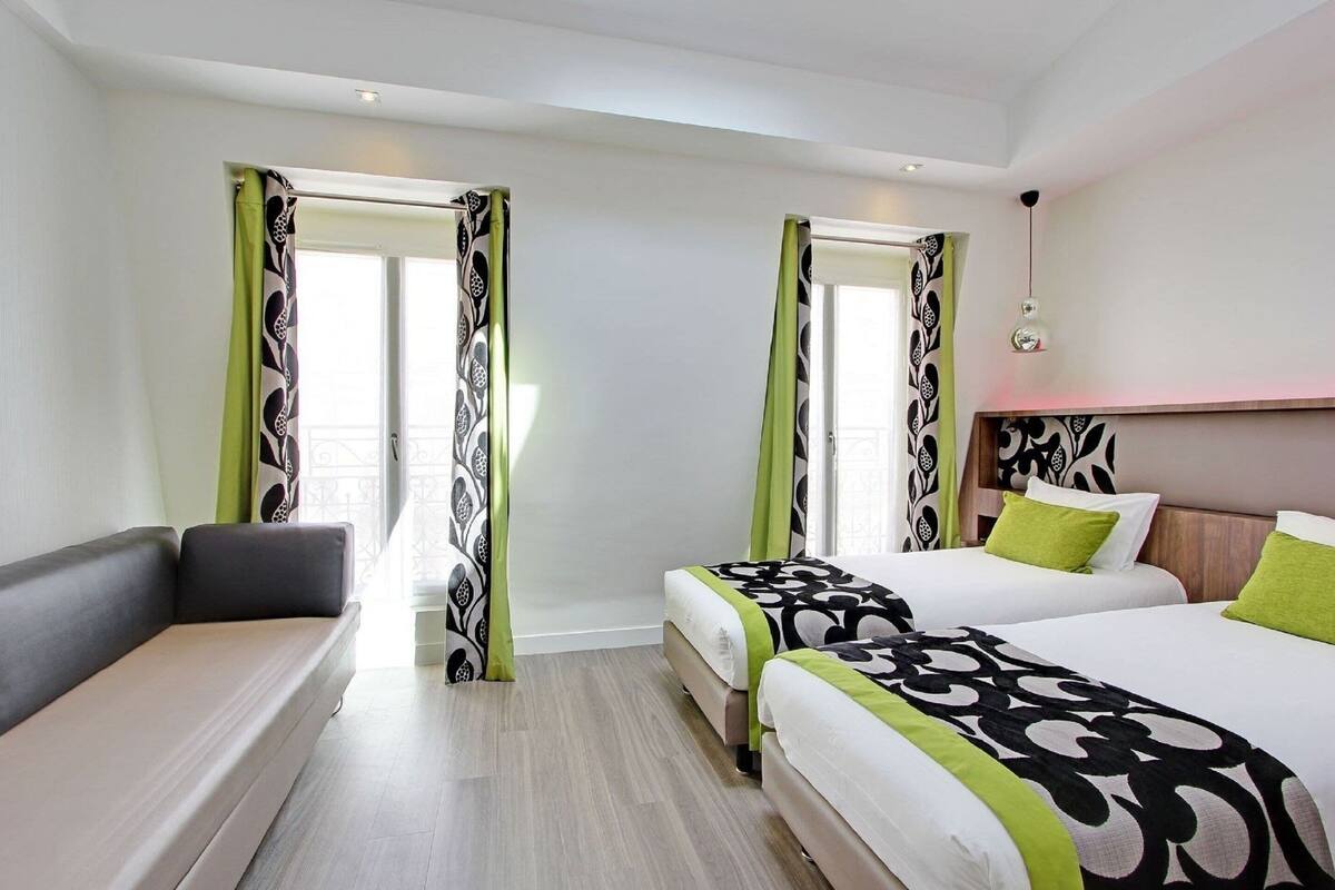 Quarto de hotel com duas camas, um sofá e uma janela grande com cortinas estampadas.