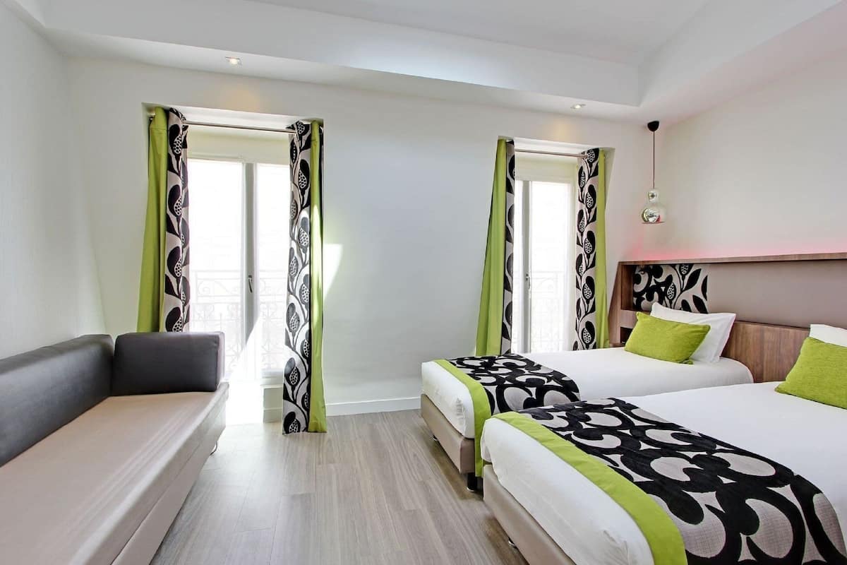 Quarto de hotel com duas camas, um sofá e uma janela grande com cortinas estampadas.