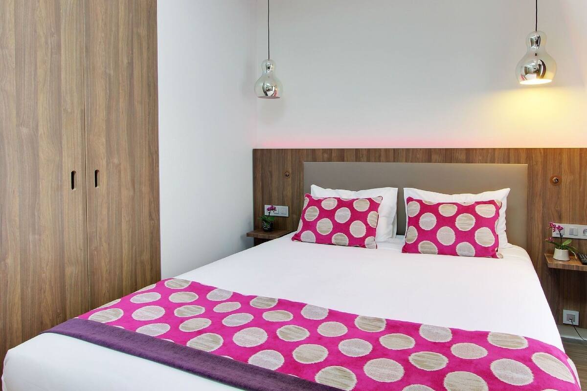 Cama bem arrumada com colcha e almofadas rosa e brancas com bolinhas, cabeceira de madeira e duas luminárias laterais.