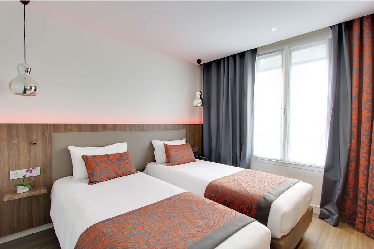 Quarto de hotel com duas camas, um encosto de madeira, uma mesinha de cabeceira com uma planta e uma janela com cortinas.