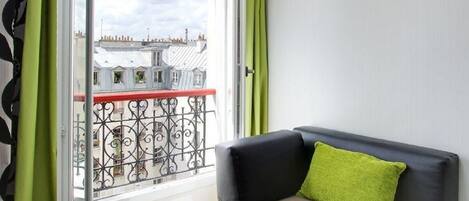A balcony with a wrought iron railing, a black sofa with a green pillow, and a view of buildings through the railing.