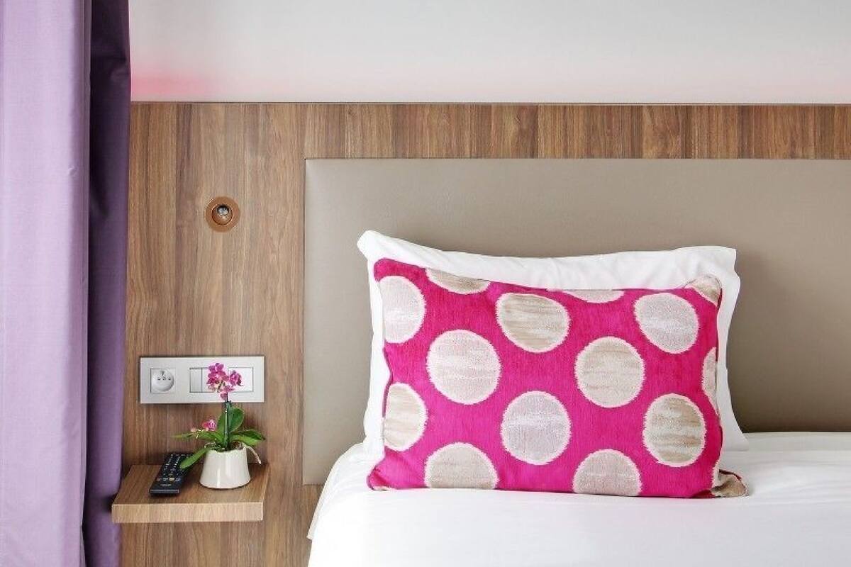Uma cama bem arrumada com um travesseiro rosa com bolinhas brancas, uma mesa de cabeceira com controle remoto e uma planta em vaso, e uma cabeceira de madeira.