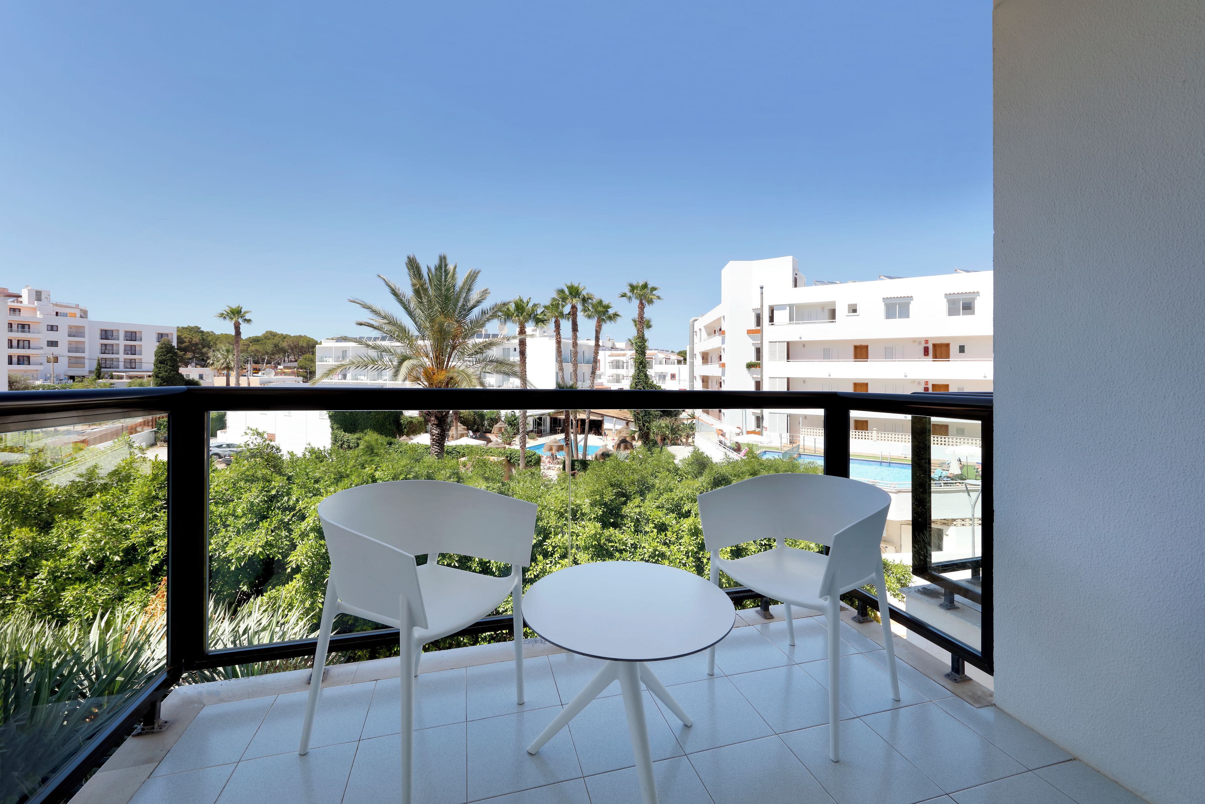 Un balcon avec deux chaises blanches et une petite table, donnant sur une piscine et des immeubles.