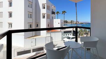 Un balcón con sillas y una mesa blancos, con vistas a una zona costera con edificios y un cielo azul despejado.