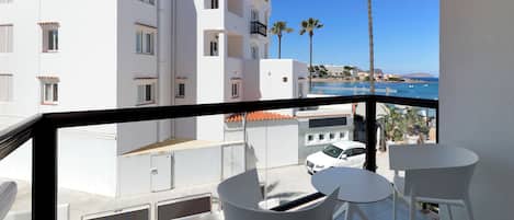 A balcony with white chairs and a table, overlooking a coastal area with buildings and a clear blue sky.
