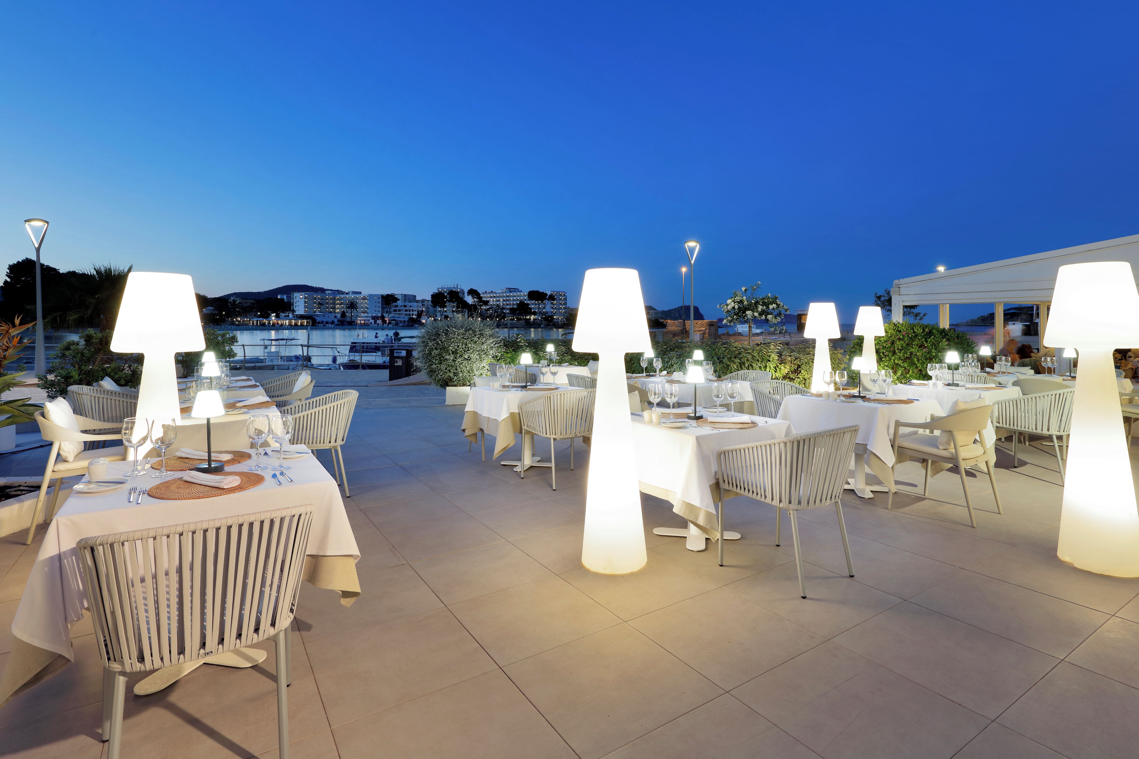 An outdoor dining area with tables and chairs, illuminated by large lamps, overlooking a body of water and buildings in the background.