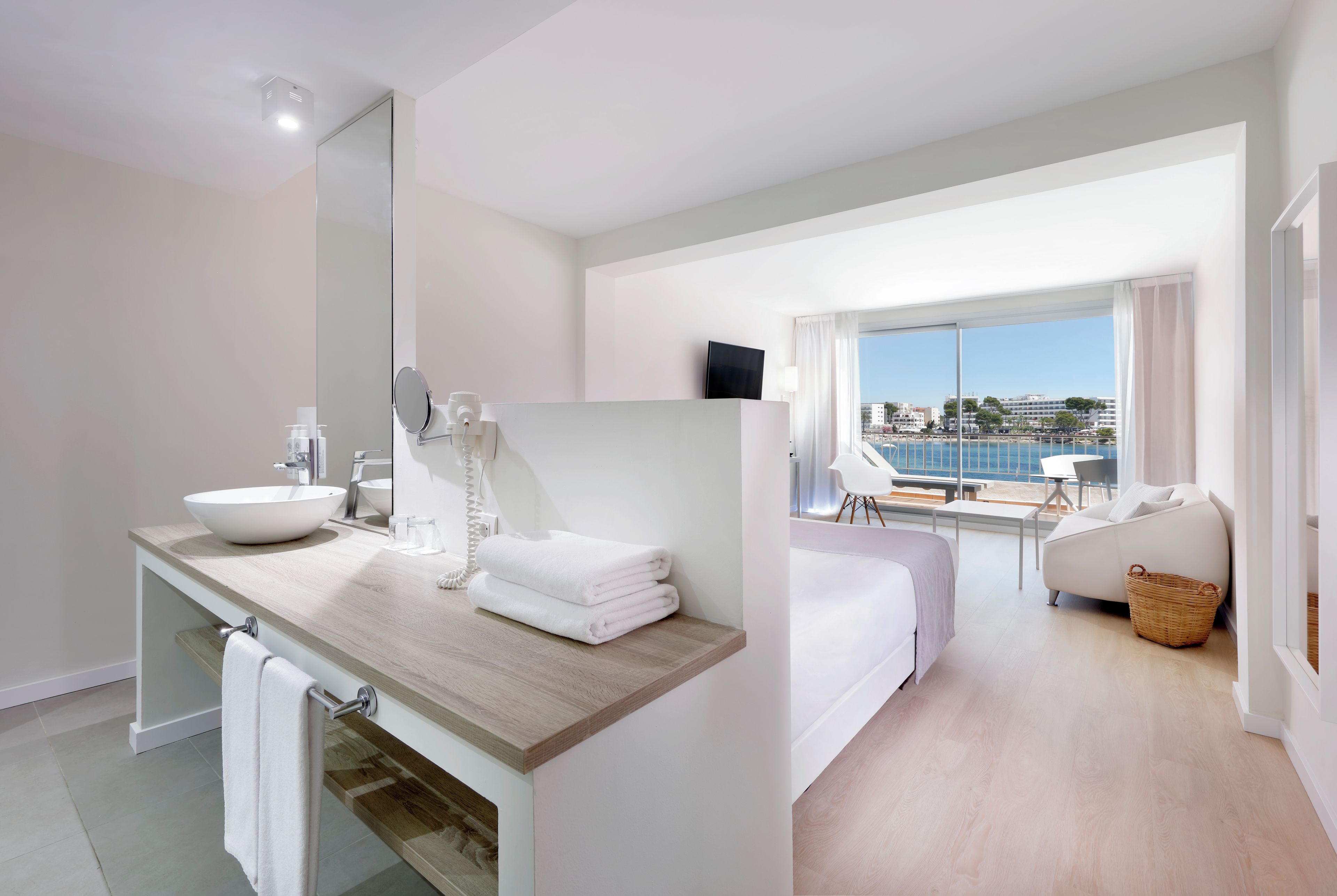 Habitación de hotel moderna con un ventanal grande, un baño con lavamanos y espejo, y un balcón con vista al mar.