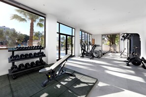 Un gimnasio moderno con equipo de ejercicio, ventanas amplias y vistas a palmeras y edificios.