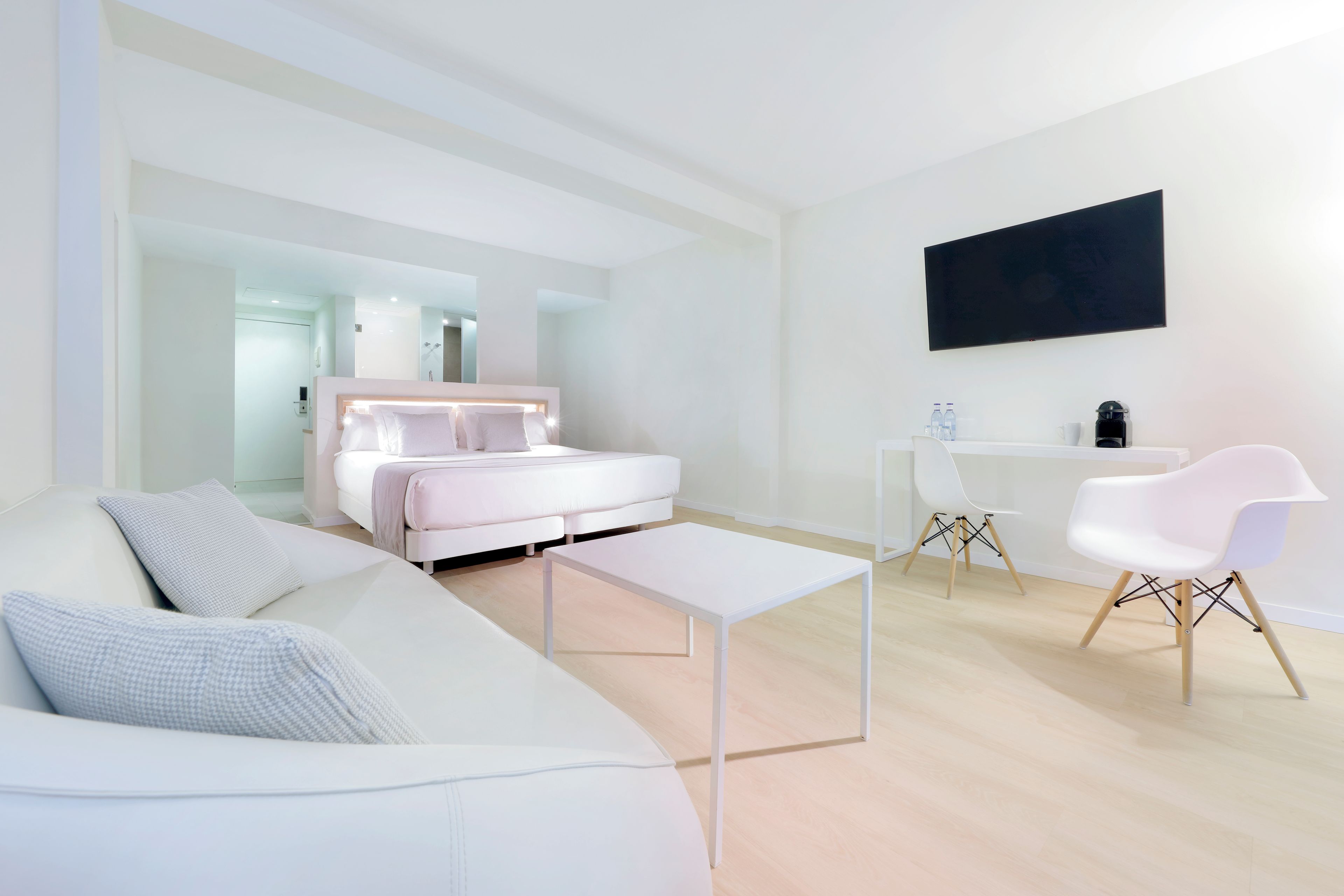 A modern hotel room with a white sofa, a bed with a headboard, a small white table, a desk with a chair, and a flat-screen TV mounted on the wall.
