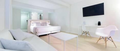Habitación de hotel moderna con un sofá blanco, una cama con cabecera, una mesita pequeña blanca, un escritorio con silla y un televisor de pantalla plana montado en la pared.