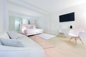 Une chambre d’hôtel moderne avec un canapé blanc, un lit doté d’une tête de lit, une petite table blanche, un bureau avec une chaise et un téléviseur à écran plat fixé au mur.