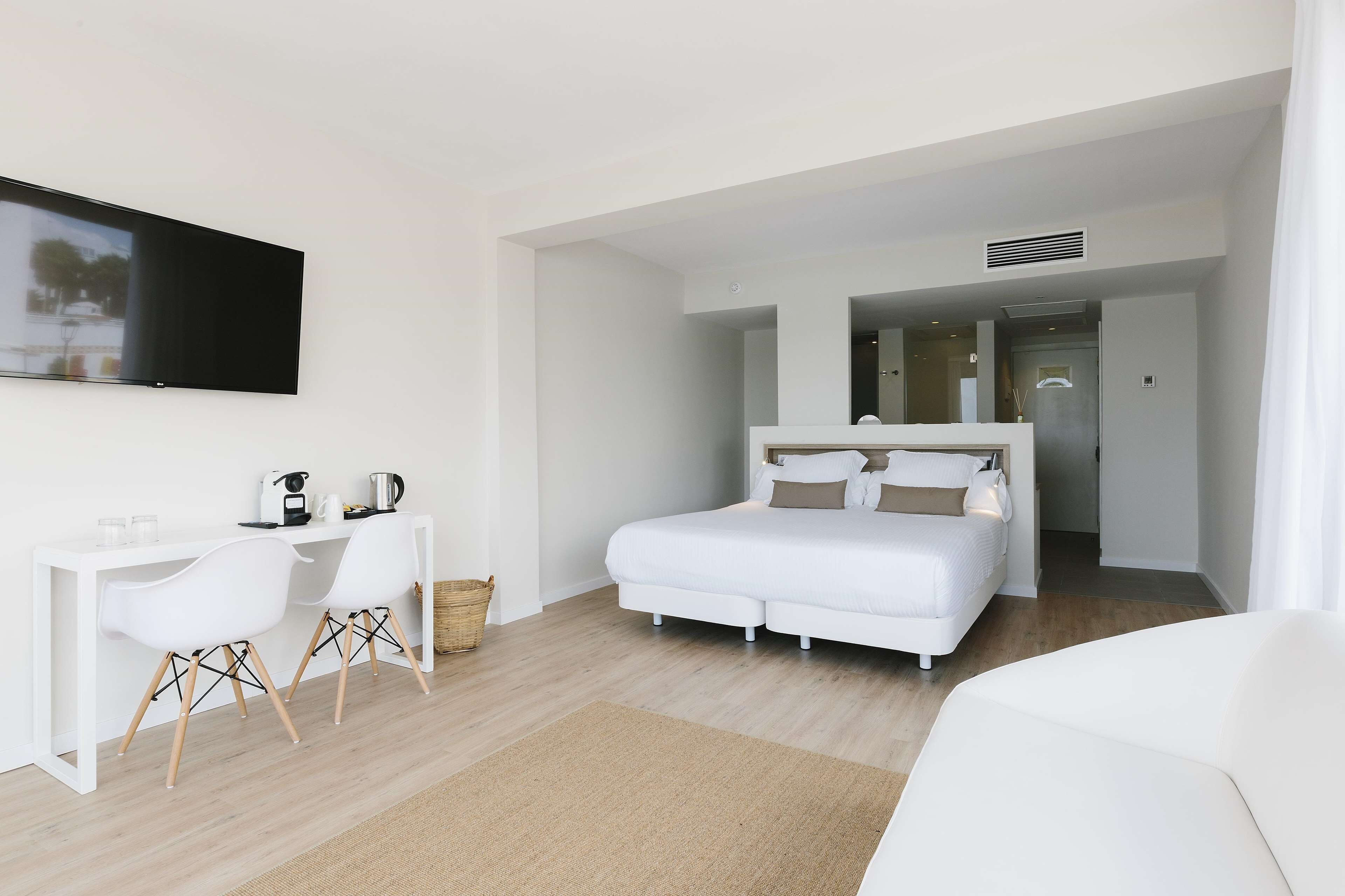 A modern hotel room with a white bed, a desk with two chairs, a flat-screen TV mounted on the wall, and a large mirror above the bed.